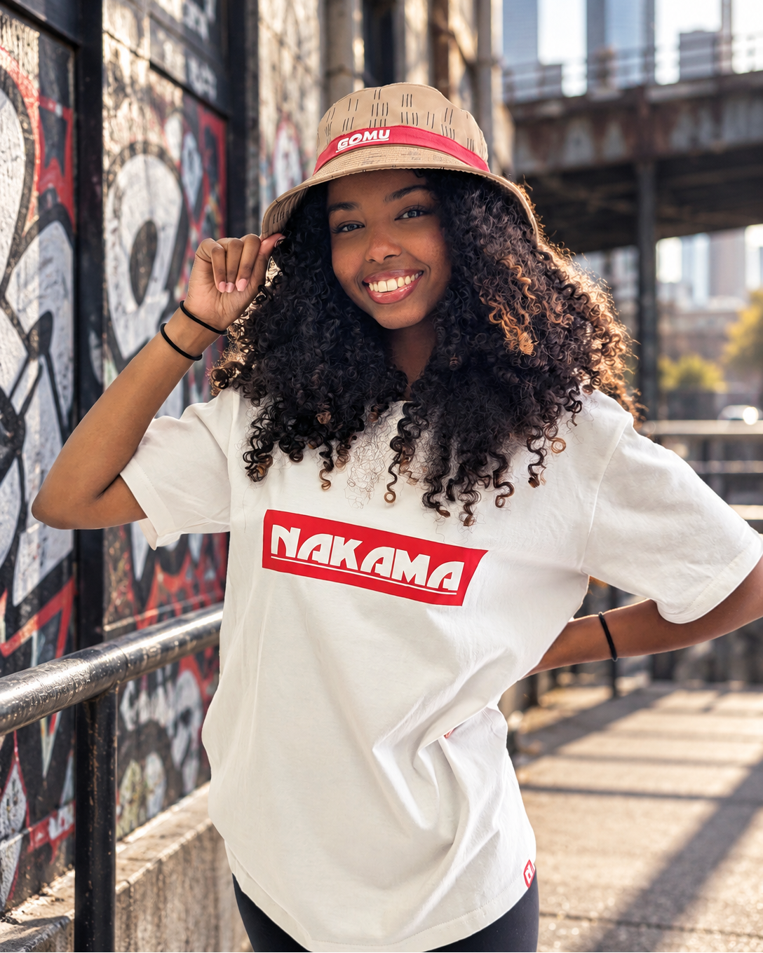 A smiling woman with curly hair stands outdoors wearing a white t-shirt with a red 'NAKAMA' box logo on the chest, a beige patterned bucket hat with a red band reading 'GOMU', and black pants. The background shows a graffiti-covered wall and an urban bridge structure.